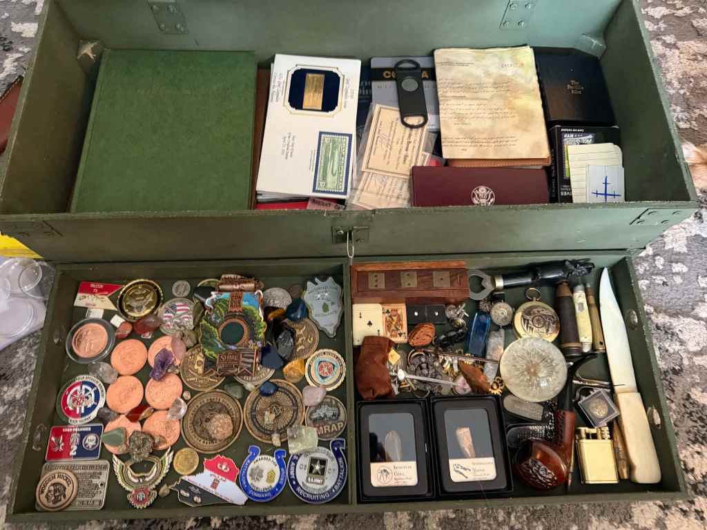 Open green military footlocker filled with challenge coins, pins, pocket knives, lighters and other keepsakes